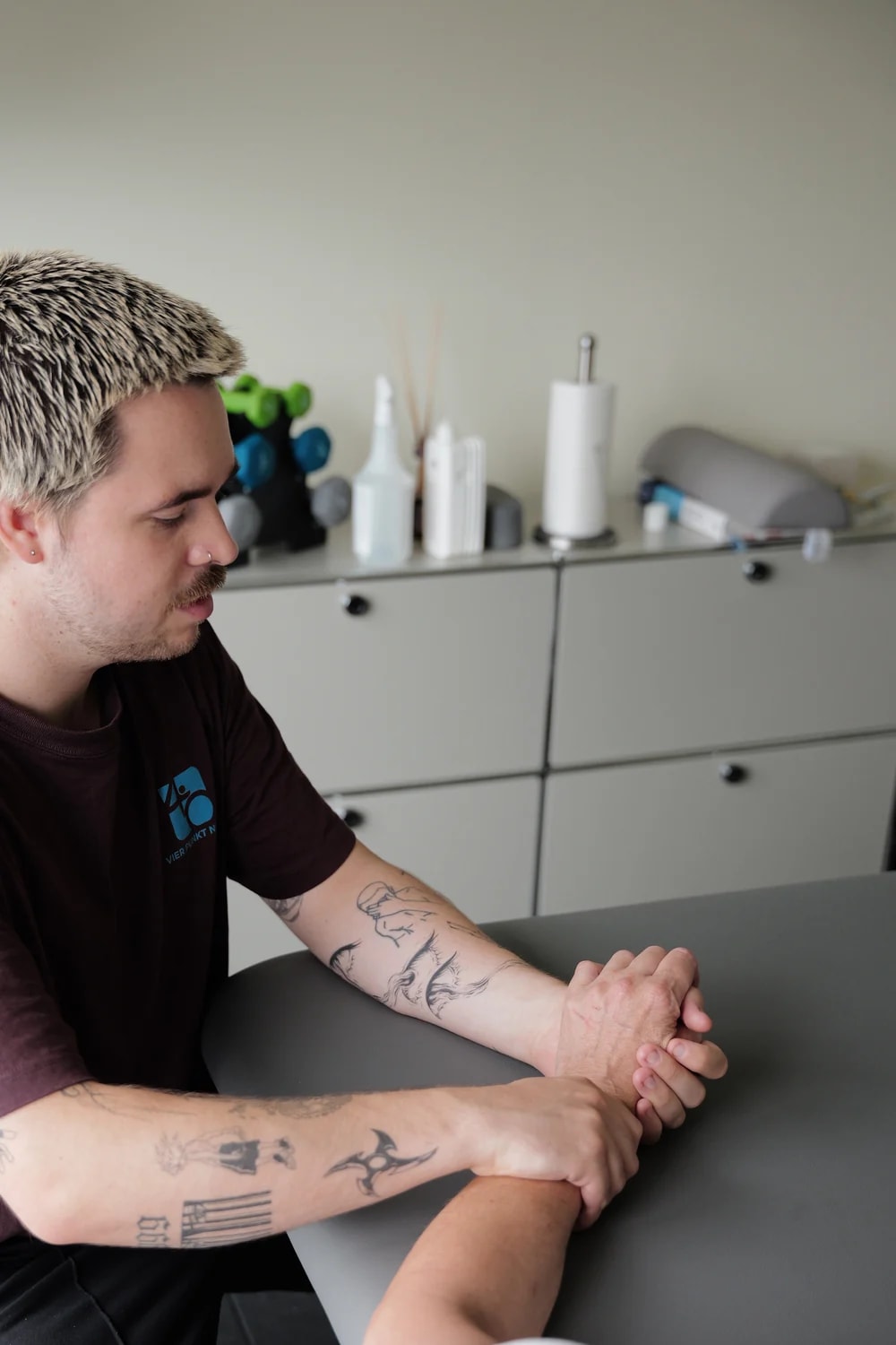 Therapeut arbeitet am Handgelenk einer Person, im Hintergrund das Materialsideboard.