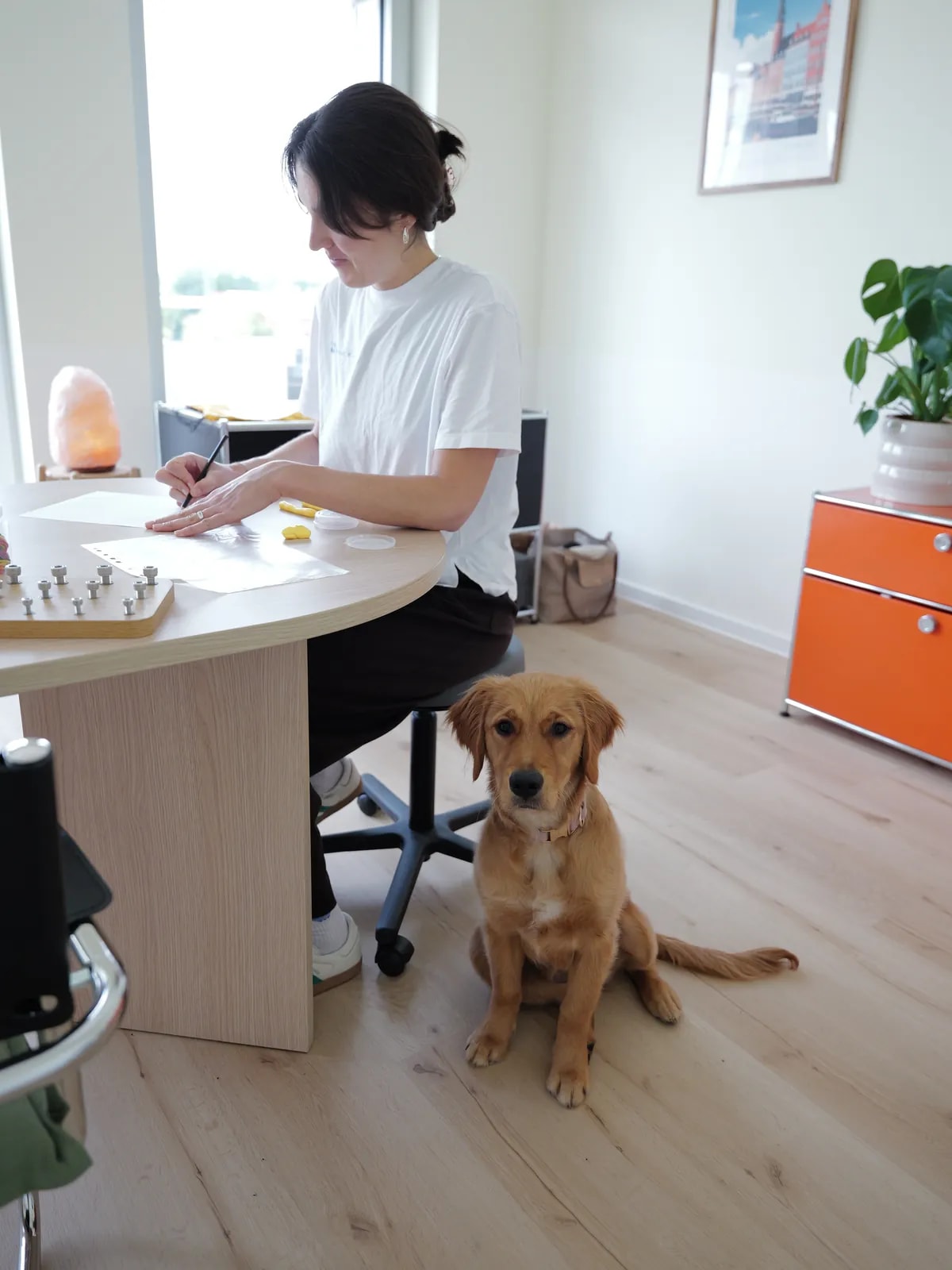 Mitarbeiterin im weißen T-Shirt schreibt an einem runden Holztisch mit Kleinmaterial, neben ihr sitzt ein junger Hund.