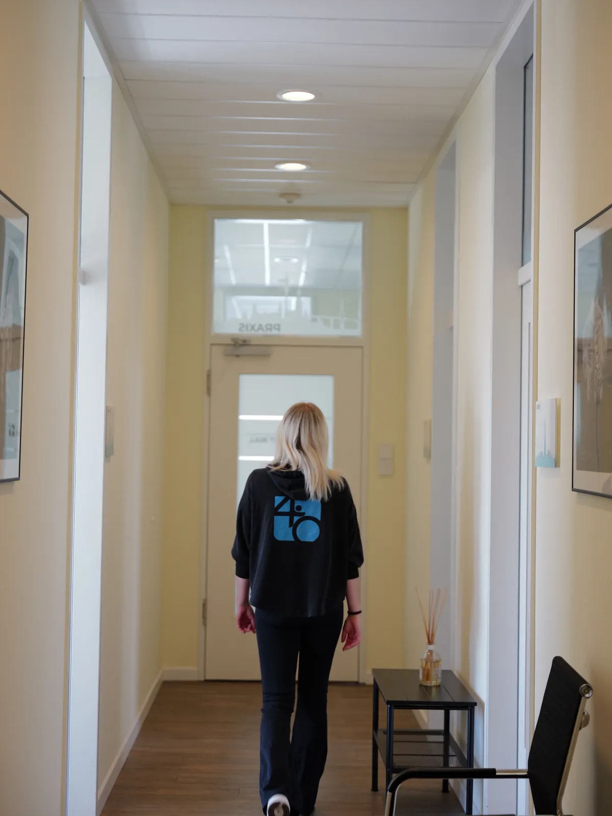 Mitarbeiterin in Praxis-Hoodie geht von hinten durch einen hellen Flur mit Holzboden auf eine Tür zu.