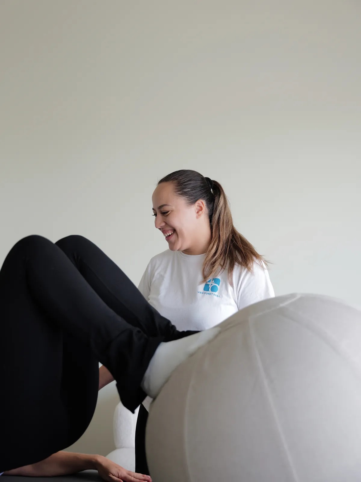 Patientin liegt auf dem Rücken, die Beine auf einem Gymnastikball, die Therapeutin lacht dahinter.