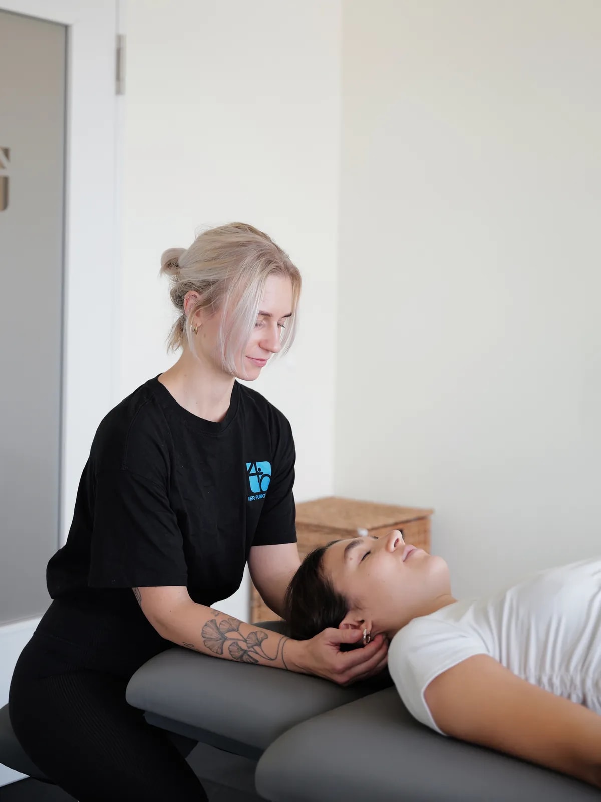 Therapeutin im schwarzen Praxis-Shirt hält mit beiden Händen den Nacken einer Patientin, die auf der Behandlungsliege liegt.