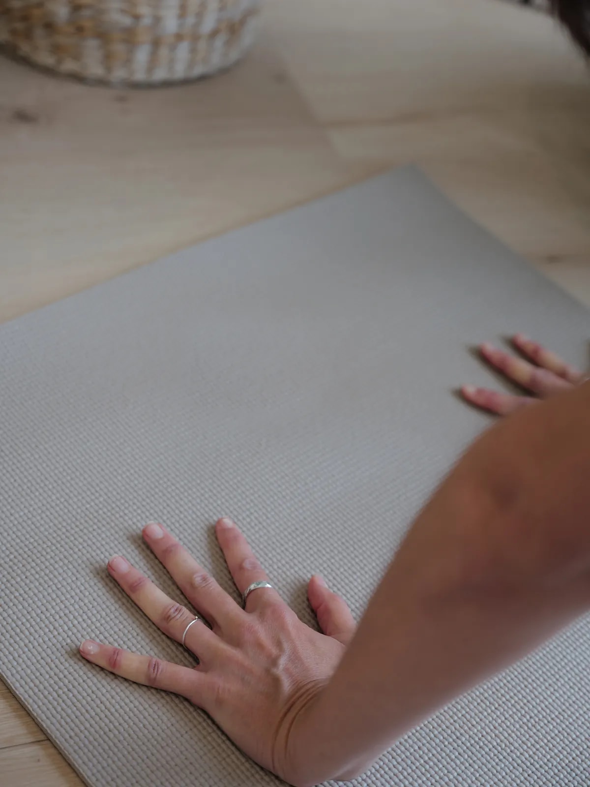 Hand mit Ringen liegt flach auf einer grauen Matte auf dem Holzboden, daneben ein Korb und ein zweiter Unterarm.