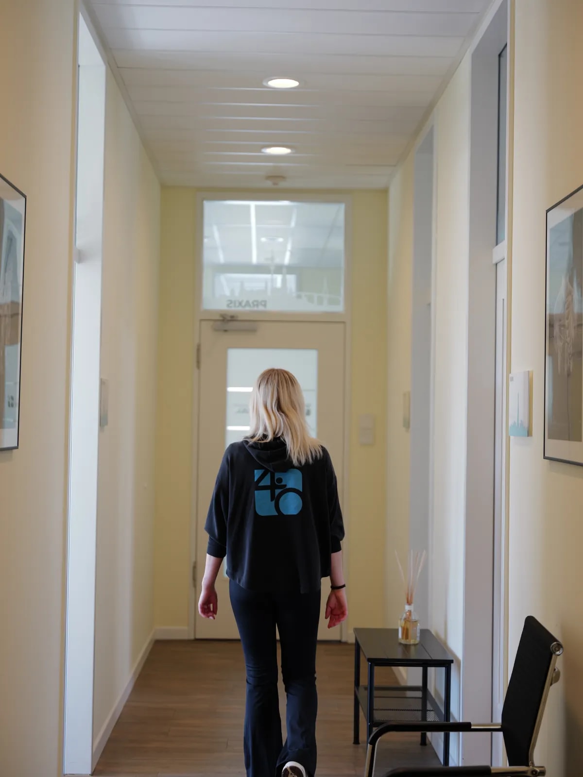 Mitarbeiterin in dunkler Jacke mit Praxislogo auf dem Rücken geht von hinten durch einen Flur auf eine Glastür zu.