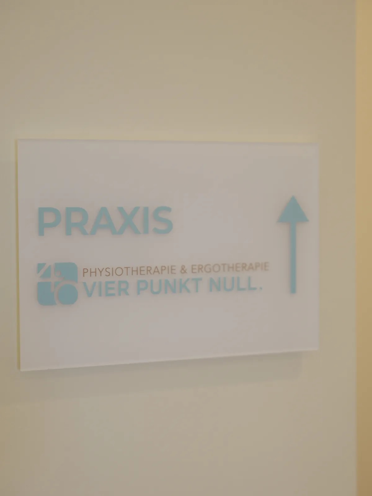 Wegweiser-Schild PRAXIS mit Pfeil nach oben und dem Logo VIER PUNKT NULL an heller Wand.