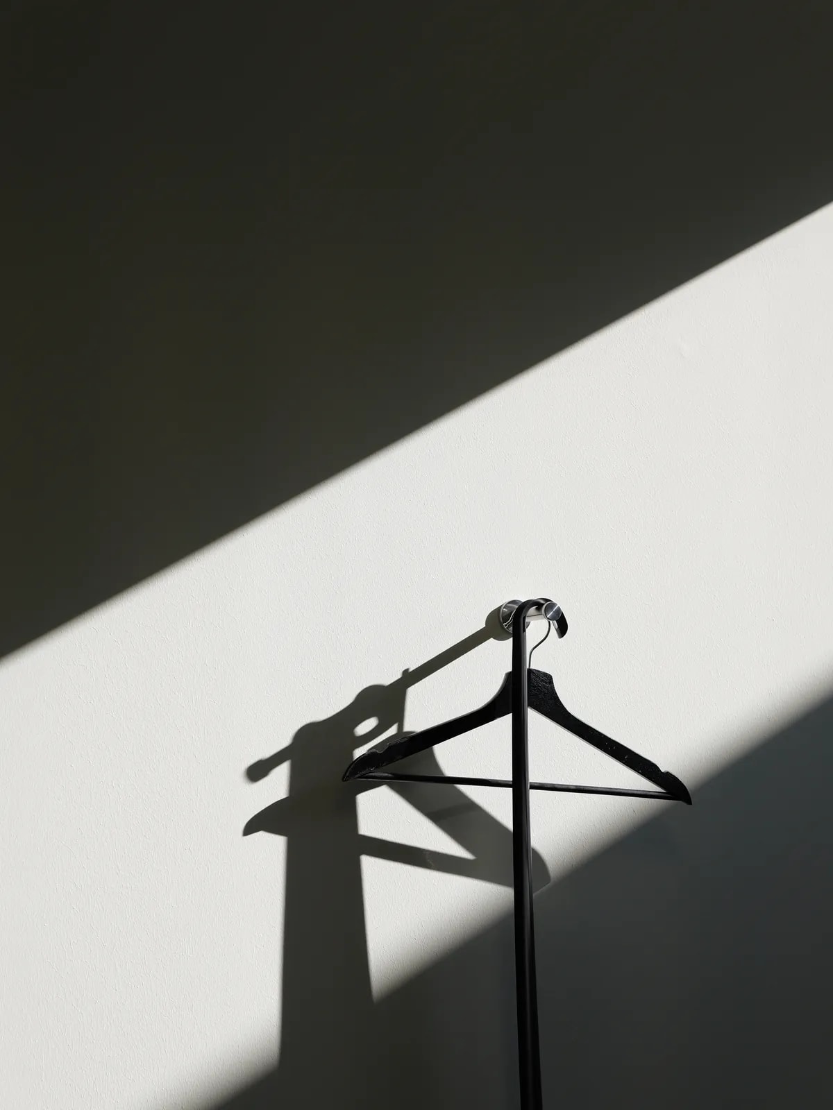 Schwarzer Kleiderbügel an einem Haken vor heller Wand, schräg fällt Sonnenlicht und wirft Schatten.