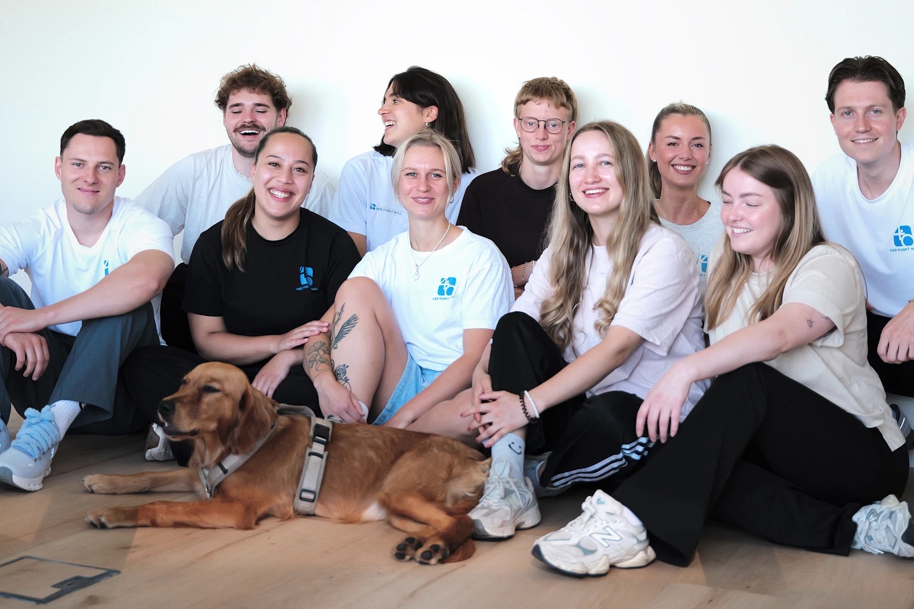 Das Team von Vier Punkt Null sitzt gemeinsam mit dem Praxishund auf dem Boden und lacht in die Kamera.